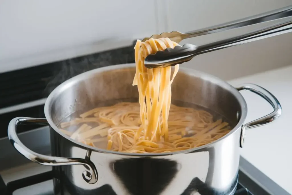 A stainless steel pot on a stove with steaming water and cooking fettuccini pasta, lifted by tongs showcasing the cooked noodles