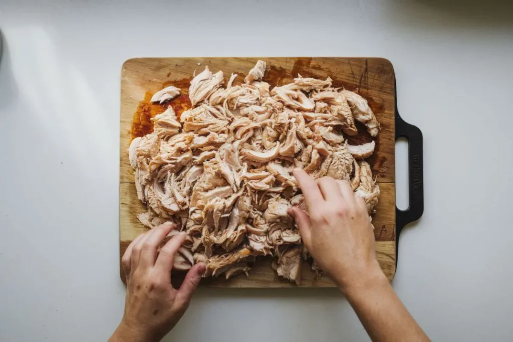 Overhead shot of a cutting board with shredded roasted chicken. Hands are touching the chicken. The board is placed over a well-lit white countertop, with a clean and simple background