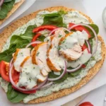 Cottage cheese flatbread topped with grilled chicken, fresh tomatoes, red peppers, onions, and mixed greens, drizzled with yogurt pesto sauce, served on a rustic wooden board