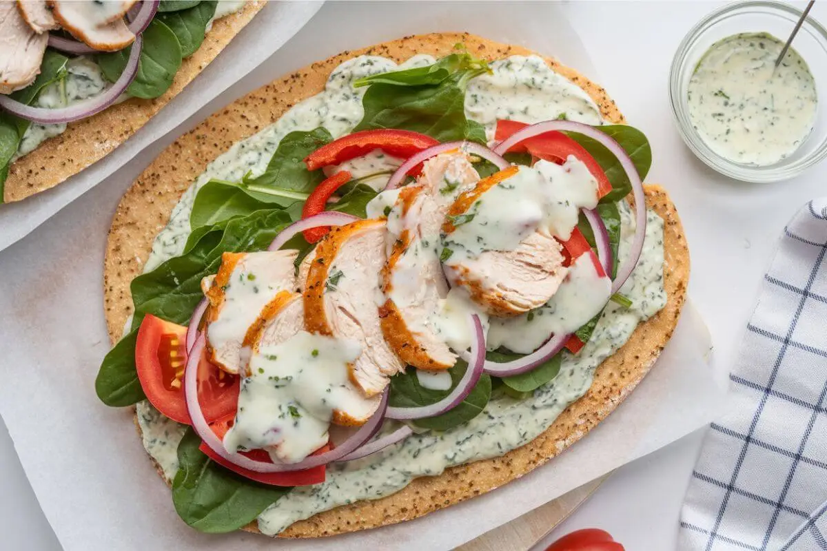 Cottage cheese flatbread topped with grilled chicken, fresh tomatoes, red peppers, onions, and mixed greens, drizzled with yogurt pesto sauce, served on a rustic wooden board