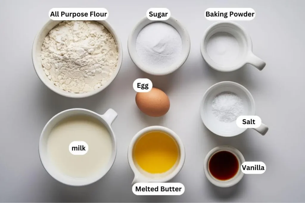 All ingredients for mini pancakes arranged on a white table, including flour, sugar, egg, milk, melted butter, vanilla, baking powder, and salt in small bowls